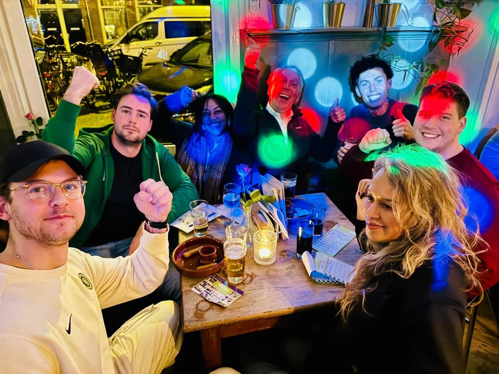 Een groep mensen viert feest in een restaurant met kleurrijke lampjes, ze maken vrolijke gebaren en lachen samen.
