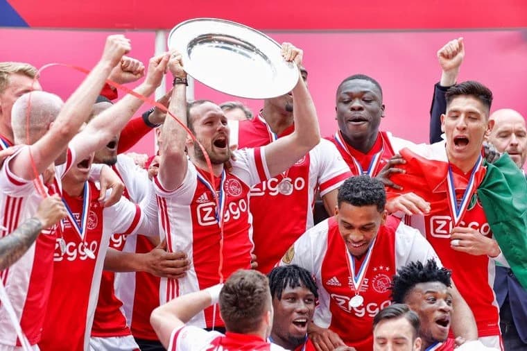 Voetbalteam viert feest met trofee en medailles, spelers in rood-wit uniformen, een speler houdt de trofee omhoog en anderen juichen, omringd door enthousiaste teamgenoten