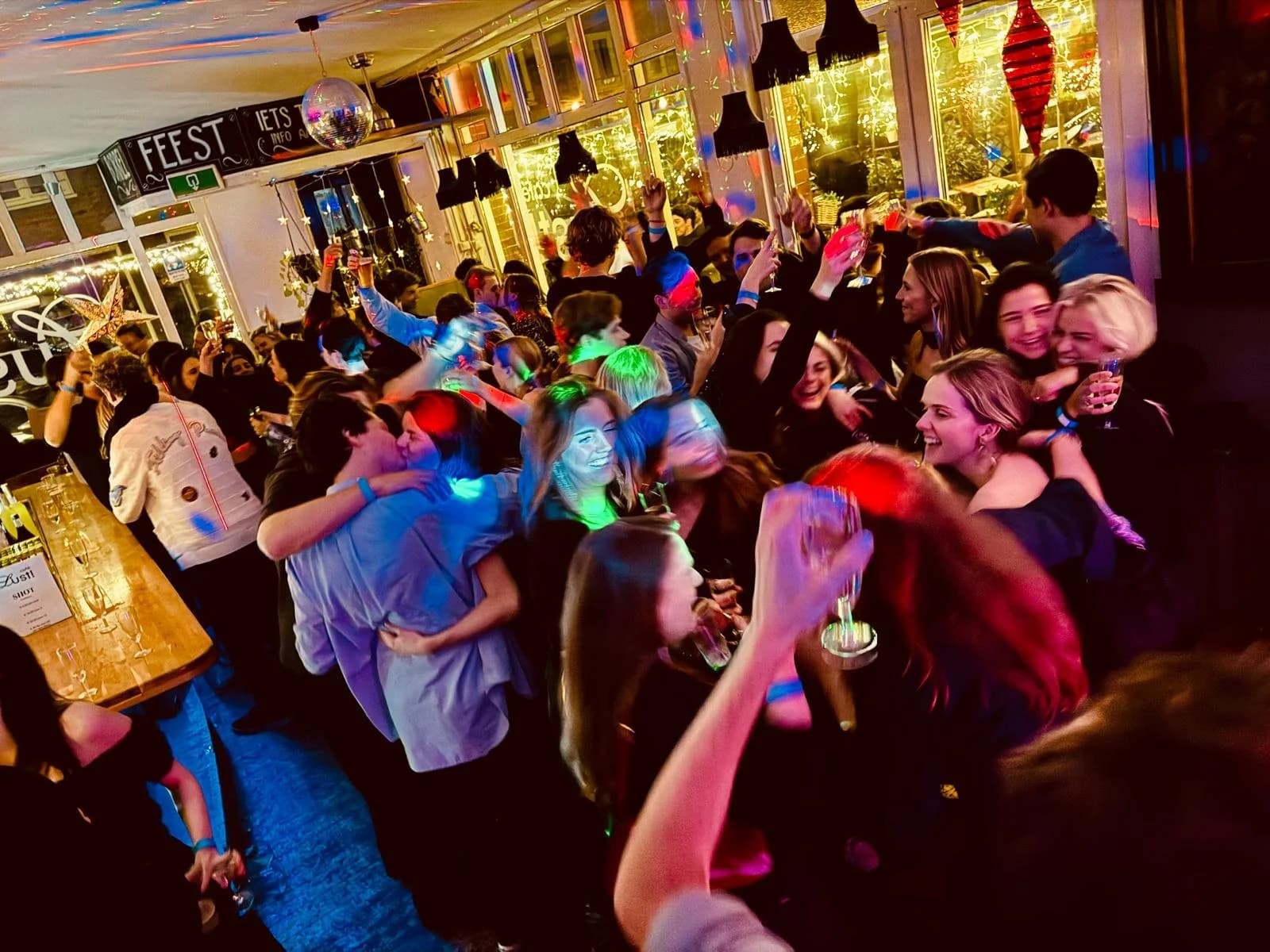 Een groep mensen die feest vieren in een gezellige binnenruimte, zij lachen, dansen en drinken. Er zijn kleurrijke lichteffecten en decoraties.